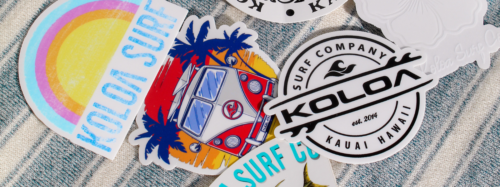 Koloa Surf Company stickers scattered across a surface. The stickers feature various colorful designs and surf-inspired graphics, all showcasing the Koloa Surf logo. The arrangement highlights the diversity and vibrancy of the sticker collection.